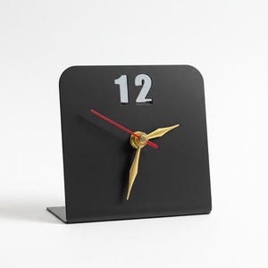 Op de afbeelding: Een L-vormige bureauklok van zwart metaal met een enkel wit cijfer 12 in het midden bovenaan. De klok heeft twee goudkleurige uur- en minutenwijzers en een dunne rode secondewijzer, gecentreerd op het matzwarte oppervlak van het paneel.