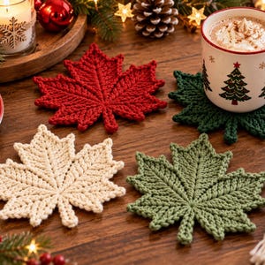 May include: A set of four crocheted maple leaf coasters in red, dark green, light green, and cream. The coasters feature detailed ribbed textures and pointed edges, arranged on a wooden surface alongside a mug and holiday decorations.