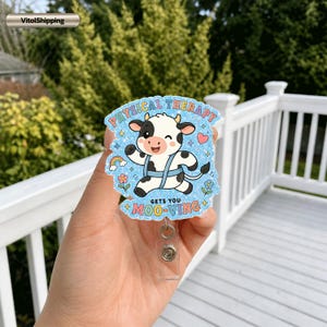 May include: A glittery badge reel featuring a cartoon cow wearing a blue harness, surrounded by a rainbow, flowers, and stars. The text reads PHYSICAL THERAPY GETS YOU MOO-VING in colorful letters. The badge has a clear plastic clip and retractable cord.