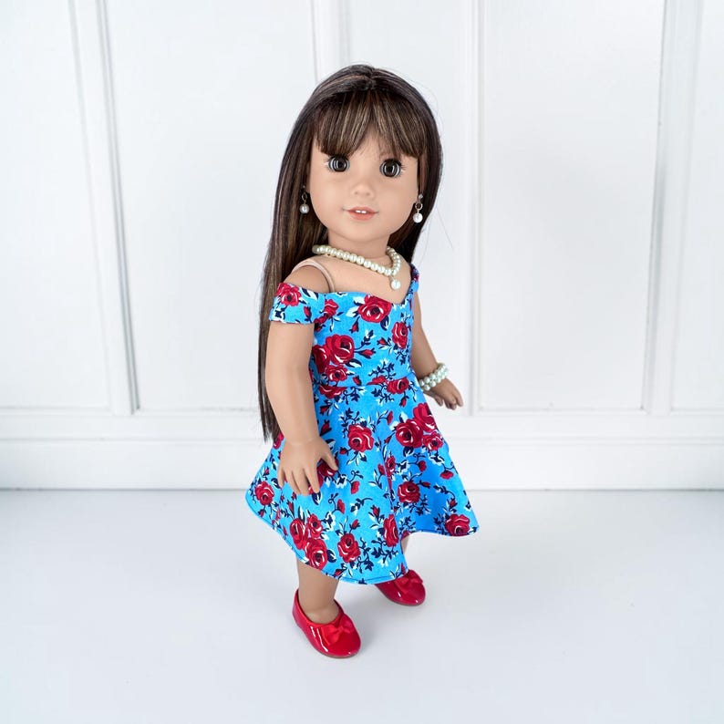 K&ouml;nnte beinhalten: Eine Puppe mit langem braunem Haar und Pony tr&auml;gt ein schulterfreies blaues Kleid mit einem roten Rosenmuster. Die Puppe ist mit einer wei&szlig;en Perlenkette, passenden Perlenohrringen, einem Perlenarmband und gl&auml;nzenden roten flachen Schuhen ausgestattet.
