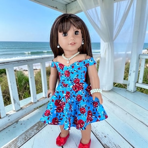 K&ouml;nnte beinhalten: Eine Puppe mit langem braunem Haar und Pony tr&auml;gt ein schulterfreies blaues Kleid mit einem roten Rosenmuster. Die Puppe ist mit einer wei&szlig;en Perlenkette, passenden Perlenohrringen, einem Perlenarmband und roten Schuhen ausgestattet.