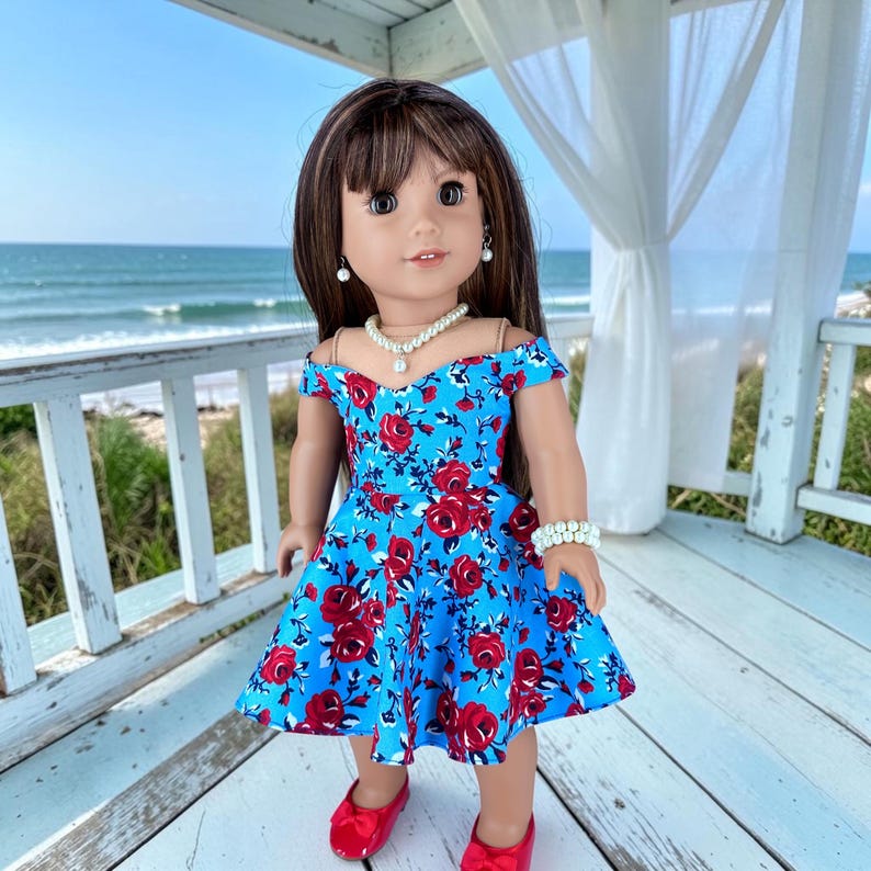 K&ouml;nnte beinhalten: Eine Puppe mit langem braunem Haar und Pony tr&auml;gt ein schulterfreies blaues Kleid mit einem roten Rosenmuster. Die Puppe ist mit einer wei&szlig;en Perlenkette, passenden Perlenohrringen, einem Perlenarmband und roten Schuhen ausgestattet.