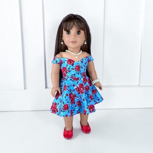 K&ouml;nnte beinhalten: Eine Puppe mit langem braunem Haar und Pony tr&auml;gt ein leuchtend blaues schulterfreies Kleid mit einem roten Rosenmuster. Die Puppe ist mit einer wei&szlig;en Perlenkette, passenden Perlenohrringen, einem Perlenarmband und roten Schuhen ausgestattet.