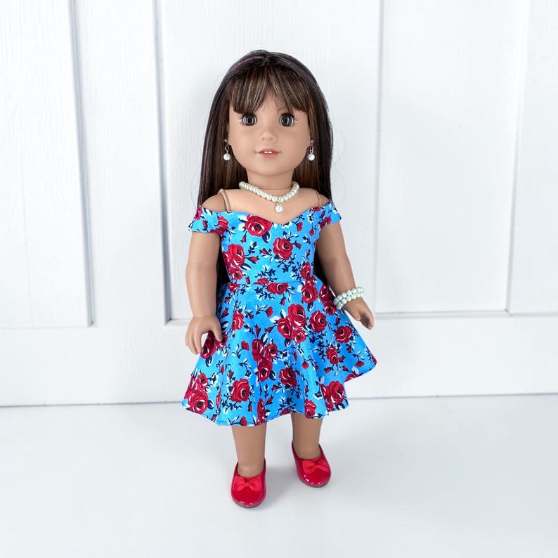 K&ouml;nnte beinhalten: Eine Puppe mit langem braunem Haar und Pony tr&auml;gt ein leuchtend blaues schulterfreies Kleid mit einem roten Rosenmuster. Die Puppe ist mit einer wei&szlig;en Perlenkette, passenden Perlenohrringen, einem Perlenarmband und roten Schuhen ausgestattet.