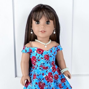 K&ouml;nnte beinhalten: Eine Puppe mit langem braunem Haar und Pony, bekleidet mit einem schulterfreien blauen Kleid mit rotem Rosenmuster. Die Puppe ist mit einer wei&szlig;en Perlenkette, passenden Ohrringen und einem Perlenarmband am linken Handgelenk geschm&uuml;ckt.