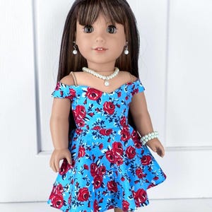 K&ouml;nnte beinhalten: Eine Puppe mit langem braunem Haar und Pony tr&auml;gt ein schulterfreies Kleid mit hellblauem Hintergrund und einem roten Rosenmuster. Die Puppe ist mit einer wei&szlig;en Perlenhalskette, passenden Ohrringen und einem Armband aus Perlen geschm&uuml;ckt.