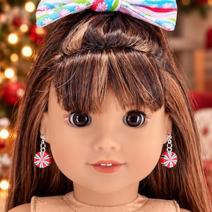 May include: A doll with long brown hair and bangs, wearing a colorful patterned hair bow and dangling earrings shaped like round red, white, and green peppermint candies. The doll has dark eyes, a neutral expression, and a small gap between front teeth.