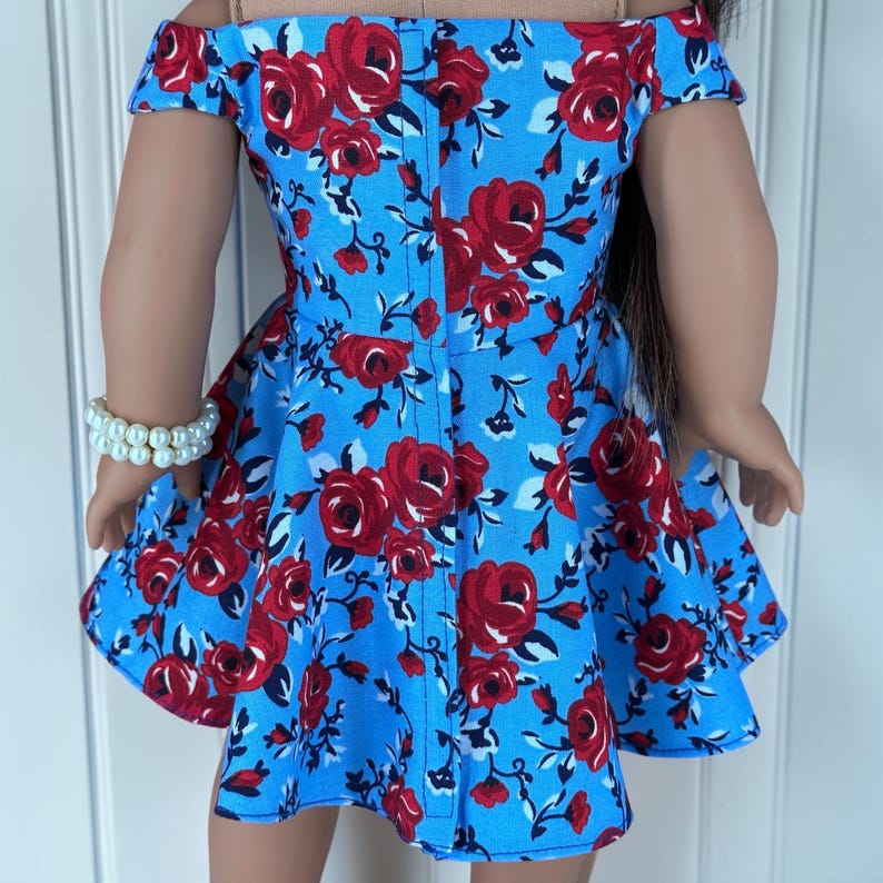 K&ouml;nnte beinhalten: Ein schulterfreies Kleid aus hellblauem Stoff mit einem sich wiederholenden Muster aus roten Rosen und dunkelgr&uuml;nen Bl&auml;ttern. Das Kleidungsst&uuml;ck hat einen ausgestellten Rock und eine vertikale Naht in der Mitte des R&uuml;ckens. Ein wei&szlig;es Perlenarmband.
