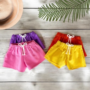 May include: Four pairs of colorful shorts in pink, purple, orange, and yellow, each with a white drawstring. The shorts are arranged on a light wood-grain surface, with a straw hat and green leaves in the background. The shorts are a summer clothing item.