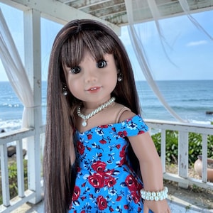 K&ouml;nnte beinhalten: Eine Puppe mit langem braunem Haar und Pony tr&auml;gt ein blaues schulterfreies Kleid mit einem roten Rosenmuster. Die Puppe ist mit einer wei&szlig;en Perlenkette, einem passenden doppelreihigen Perlenarmband und kleinen wei&szlig;en Perlenohrsteckern geschm&uuml;ckt.