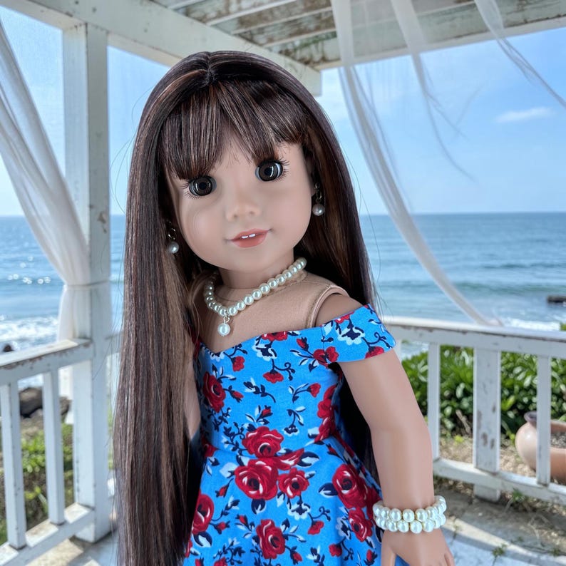 K&ouml;nnte beinhalten: Eine Puppe mit langem braunem Haar und Pony tr&auml;gt ein blaues schulterfreies Kleid mit einem roten Rosenmuster. Die Puppe ist mit einer wei&szlig;en Perlenkette, einem passenden doppelreihigen Perlenarmband und kleinen wei&szlig;en Perlenohrsteckern geschm&uuml;ckt.