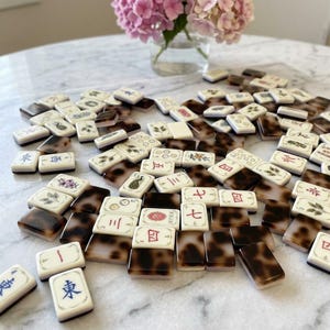 Puede incluir: Una colección de fichas de Mahjong rectangulares esparcidas sobre una superficie. Cada ficha presenta una cara blanca con varios caracteres chinos, números e ilustraciones florales coloridos, mientras que el reverso muestra un patrón de carey marrón.