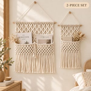 May include: A 2-piece set of off-white macrame wall hangings featuring woven cord patterns, wooden dowels, and hanging strings. The larger piece has two pockets holding The Kinfolk Home and Cereal magazines, while the smaller holds dried flowers.