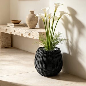 May include: A black woven round planter containing a bouquet of white calla lilies with long green stems and delicate feathery green foliage. The planter has a textured, ribbed exterior surface and sits on a light-coloured tiled floor.