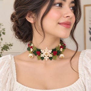 May include: A crochet choker necklace featuring a central cream-colored snowflake, dark green leaves, clusters of small red berries, and two small cream-colored bells hanging from the sides, all crafted with intricate yarn detailing.