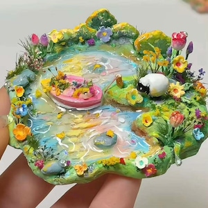 Könnte beinhalten: Ein Miniatur-Diorama mit einem Teich, einem rosa Boot voller Blumen, einem kleinen weißen Schaf mit schwarzem Gesicht und bunten, geformten Blumen an den grasbewachsenen Ufern. Die Wasseroberfläche hat eine glänzende, strukturierte Oberfläche in Blau- und Gelbtönen.