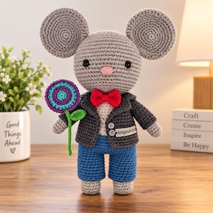 May include: A gray crocheted mouse doll with large round ears, a pink nose, and black eyes. It wears a dark gray jacket with two white buttons, a bright red bow tie, and blue pants. The mouse holds a purple and teal crocheted flower.