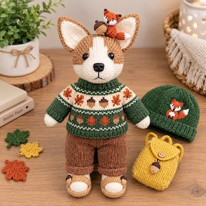 Könnte beinhalten: Ein gehäkeltes Corgi-Stofftier in einem grünen Pullover mit Eichel- und Blattmuster, brauner Cordhose und kleinen Schuhen. Ein winziger Fuchs sitzt auf dem Kopf. Zubehör umfasst eine grüne Strickmütze mit Fuchsmotiv und einen gelben Rucksack.