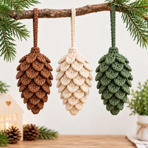May include: Three crocheted pinecone ornaments hanging from a branch. The ornaments are brown, cream, and dark green, each featuring a textured, layered scale design and a matching braided loop for hanging. The items are crafted with yarn.