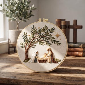 Könnte beinhalten: Ein runder Stickrahmen mit einer handgestickten Krippenszene auf weißem Stoff. Das Motiv zeigt zwei Figuren an einer Krippe unter einem großen Olivenbaum mit grünen Blättern, goldenen Sternen und einer kleinen leuchtenden Laterne.