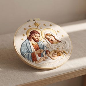 May include: A circular embroidery hoop containing a fabric panel with a detailed thread-stitched depiction of a family. The design features a bearded figure in a blue robe, a figure in a white veil, and a small infant, all with gold halos and stars.