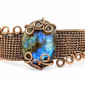 May include: A copper wire-wrapped cuff bracelet featuring a large, oval-shaped labradorite gemstone with iridescent blue and green flashes. The band is constructed from woven copper wire with decorative coiled wire accents at the edges.