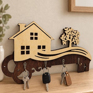 May include: A wooden key holder shaped like a large skeleton key with a light-colored house and tree cutout attached to the top. The dark brown base features four metal hooks, each holding a set of keys, including one with a brown leather tag.