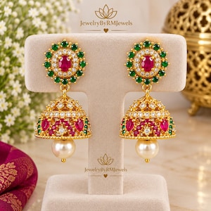 Puede incluir: Pendientes jhumka dorados con una piedra central ovalada color rubí rodeada por un halo de cristales verdes y transparentes. La base en forma de campana está adornada con piedras color rubí en forma de lágrima, cristales verdes y una perla blanca.