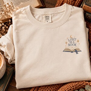 Puede incluir: Una camiseta de cuello redondo color crema con un pequeño gráfico bordado en el pecho izquierdo. El diseño muestra un libro abierto con el texto "LOST IN A BOOK" encima, rodeado de estrellas doradas y una luna creciente. Etiqueta de talla M.