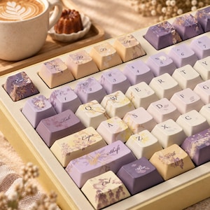 May include: A mechanical keyboard featuring a color palette of soft purple, cream, and pale yellow. The keycaps are decorated with floral patterns and botanical illustrations. The layout includes standard QWERTY lettering and function keys.