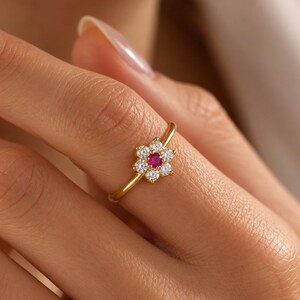 May include: A gold ring featuring a flower-shaped cluster of six clear white stones surrounding a single vibrant red stone in the center. The band is thin and polished gold, creating a delicate look with the floral arrangement on the front.
