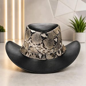Può includere: Un cappello nero con tesa larga e corona alta caratterizzato da una fascia con motivo a pelle di serpente. La fascia è accentuata da piccoli occhielli metallici attorno alla base. La parte superiore della corona è nera, abbinata alla tesa curva.