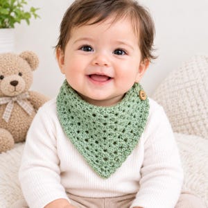 May include: A sage green triangular crochet bib featuring a textured openwork pattern and a single wooden button closure at the side. The bib is worn around the neck over a white long-sleeved shirt, showcasing a detailed yarn stitch design.