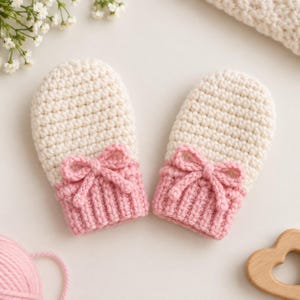 Puede incluir: Un par de manoplas tejidas a ganchillo a mano con una sección superior de color crema y un puño acanalado rosa. Cada manopla está decorada con un pequeño lazo de ganchillo rosa a juego centrado en la muñeca, con textura de hilo.