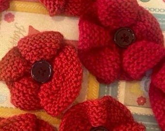 May include: A collection of five handmade red knitted flower appliques, each featuring a dark brown four-hole button at the centre. A yellow measuring tape is visible along the top edge, showing a measurement of approximately 6 centimetres.