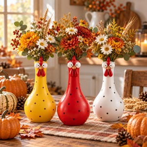Può includere: Tre vasi in ceramica a forma di gallina, colorati in giallo, rosso e bianco con pois neri. Ogni vaso presenta una testa di gallina scolpita con occhi, becco e bargigli rossi, ed è riempito con un bouquet di fiori a tema autunnale.
