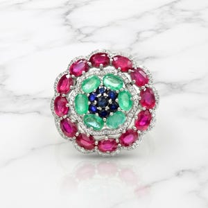 May include: A floral-shaped ring featuring a central cluster of dark blue round stones, surrounded by a ring of oval green stones, and an outer border of oval red stones, all set in silver-toned metal with small clear accent stones.