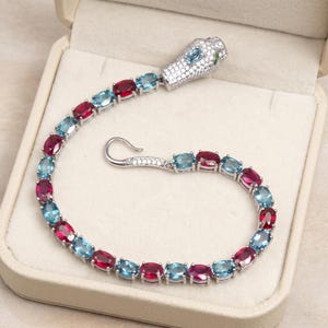 May include: A silver-toned snake bracelet featuring a pavé-set head with blue and green eyes, connected to a flexible chain of alternating oval-cut red and light blue gemstones, finished with a hook clasp and small clear stones.