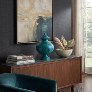 May include: A teal ceramic lidded jar with a pedestal base, positioned on a wooden sideboard next to a bowl of dried botanicals, a stack of books, and a framed abstract painting featuring gold, white, and dark blue tones.