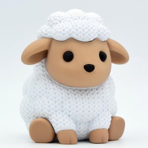 May include: A small figurine of a sitting sheep with a textured white wool body and a smooth tan face. The sheep has two large black eyes, a small black nose, and two tan ears extending from the sides of its head. The wool texture is detailed.