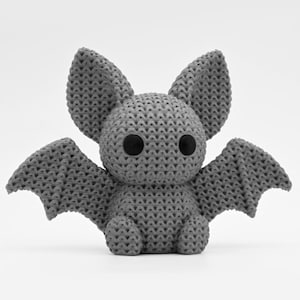 May include: A small, grey, three-dimensional bat figurine with a textured, knit-like surface pattern. The bat features two large, pointed ears, outstretched wings, and two smooth, round, solid black eyes positioned on its rounded head.