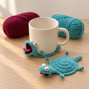 Puede incluir: Una taza de cerámica blanca con una funda de ganchillo color verde azulado en forma de rana que envuelve la base, con la boca abierta y una lengua rosa. Al lado hay un posavasos de ganchillo a juego con forma de rana y ojos blancos.