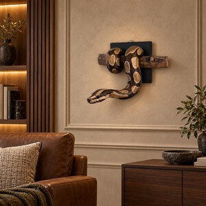 May include: A wall-mounted sculpture featuring a realistic brown and tan patterned snake coiled around a horizontal wooden branch. The branch is attached to a square black backing plate, creating a three-dimensional display piece.
