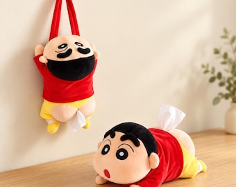 Puede incluir: Dos portapañuelos de peluche con forma de personaje de dibujos animados con pelo negro, camiseta roja y pantalones cortos amarillos. Uno cuelga de un gancho de pared con una correa roja, mientras el otro yace plano con un pañuelo blanco saliendo.