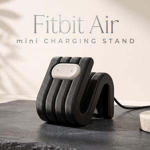 Könnte beinhalten: Ein schwarzer, gerippter, S-förmiger Ladeständer für ein Fitbit Air Gerät. Der Ständer verfügt über eine hellgraue, ovale Ladeschnittstelle auf der Vorderseite. Der Text "Fitbit Air mini CHARGING STAND" ist im Hintergrund über dem Objekt zu sehen.