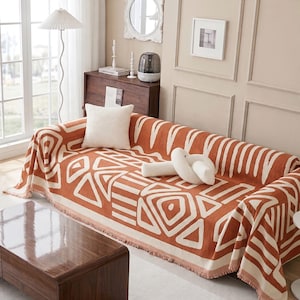 May include: A rectangular sofa throw blanket featuring a burnt orange and cream geometric pattern with fringe edges, draped over a sofa with a white square pillow and a white knotted decorative pillow placed on top.