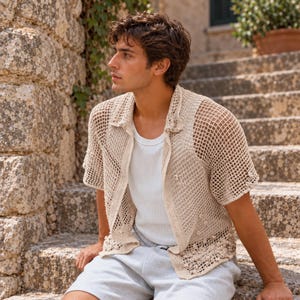 May include: A beige short-sleeved button-down shirt featuring an open-knit crochet construction with a textured grid pattern and small decorative bobble accents along the hem and front, worn over a plain white ribbed tank top and light gray shorts.