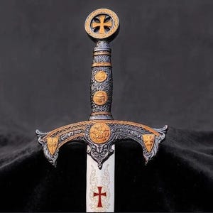Op de afbeelding: Een decoratief zwaard met een zilver- en goudkleurig gevest met ingewikkelde reliëfdetails. De pommel toont een kruis, terwijl de pareerstang gouden accenten in cirkel- en schildvorm bevat. Het lemmet heeft een geëtst rood kruisontwerp.