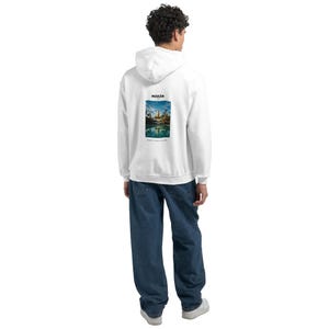 May include: A white hooded sweatshirt featuring a rectangular graphic on the back. The graphic displays a landscape with a golden structure, palm trees, and a reflection in water, with the text MAKAN above and MORE THAN A PLACE below.
