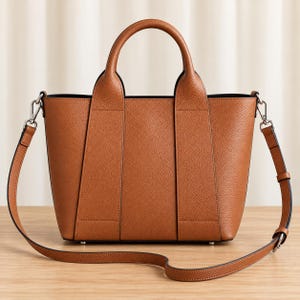 Può includere: Una borsa a mano strutturata color cuoio con texture granulata, dotata di due manici superiori e una tracolla rimovibile e regolabile. La borsa presenta una silhouette trapezoidale con dettagli a pannelli verticali e clip in metallo argentato.